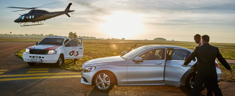 A Close Protection Officer holding a car door for an executive businessman. There is a G4S branded vehicle on the runway. A non-branded helicopter is flying off over the G4S-branded vehicle.